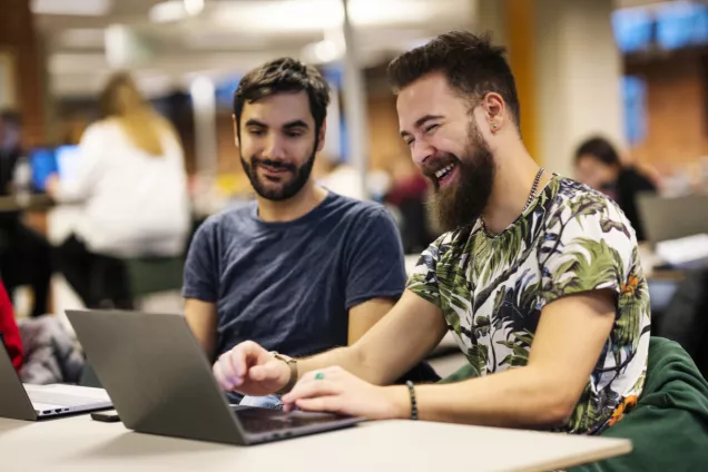 Two male students look at a computer and laugh. Photo.