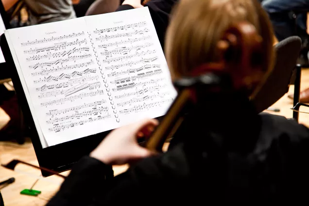 Student playing a cello in front of note sheets. Photo.