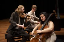 Tre musiker två män och en kvinna sitter och spelar på cello piano och violin