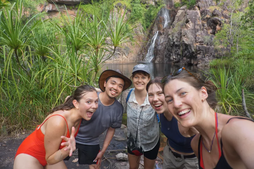 Fem studenter ler och tittar in i kameran framför ett vattenfall. Foto. 