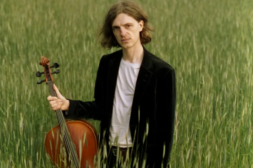Aramis Silvereke står med en cello i handen i en fält. Foto.